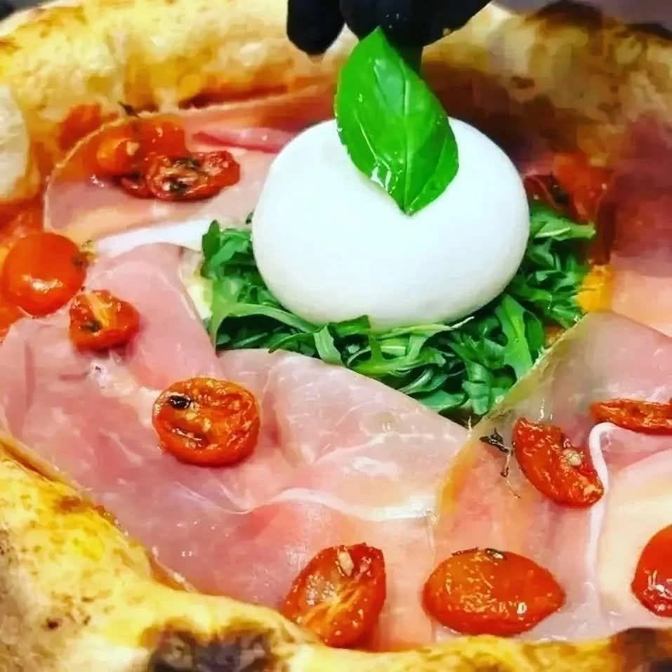 Pizza Ai Navigli con prosciutto crudo, pomodorini arrostiti, rucola e burrata su cui una mano guantata posa il basilico