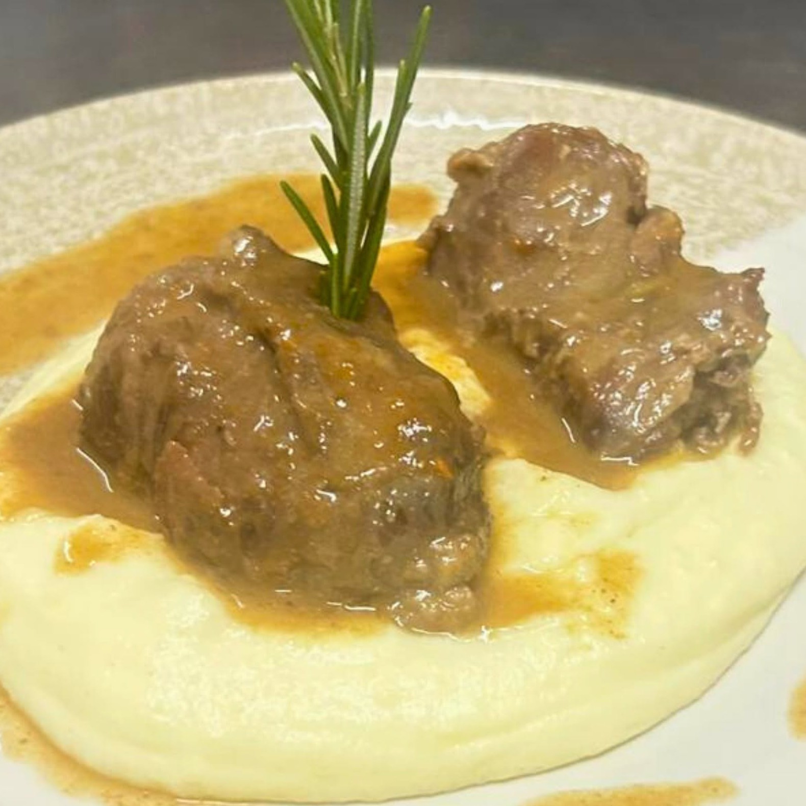 Due pezzi di carne brasata con sugo bruno su purè cremoso e rametto di rosmarino, in piatto chiaro di Ai Navigli